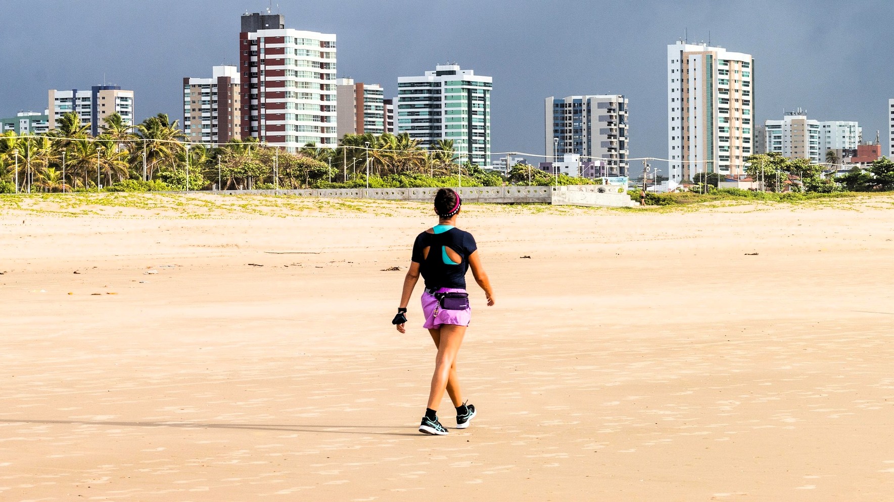 Mujer caminando conscientemente por una playa amplia con la ciudad al fondo.